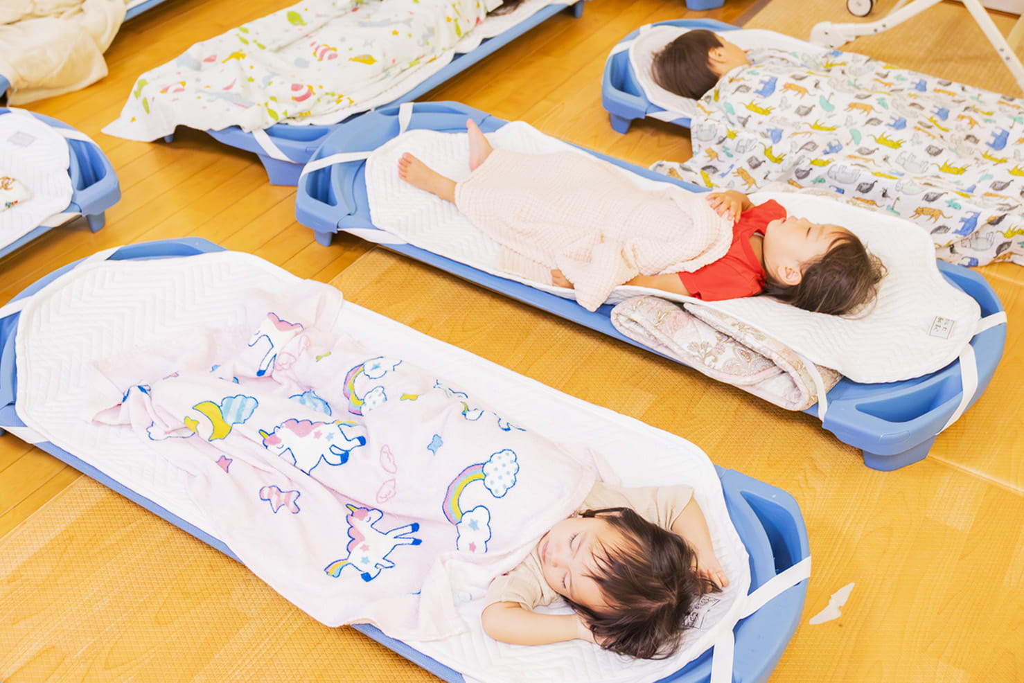 保育園の昼寝タイムに、子どもたちが簡易ベッドで毛布にくるまって寝ている様子