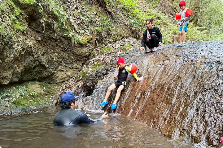 滝のような斜面を子どもが滑り降り、大人が水中で受け止めている川遊びの様子