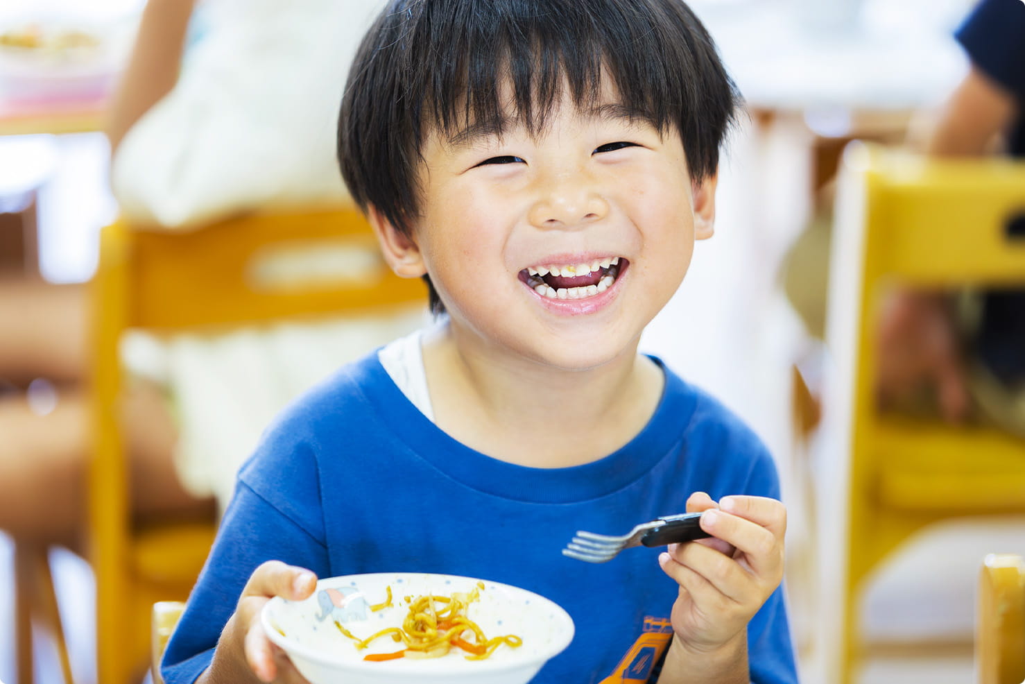 青い服を着た男の子が給食を食べながら笑顔を見せている保育園の様子
