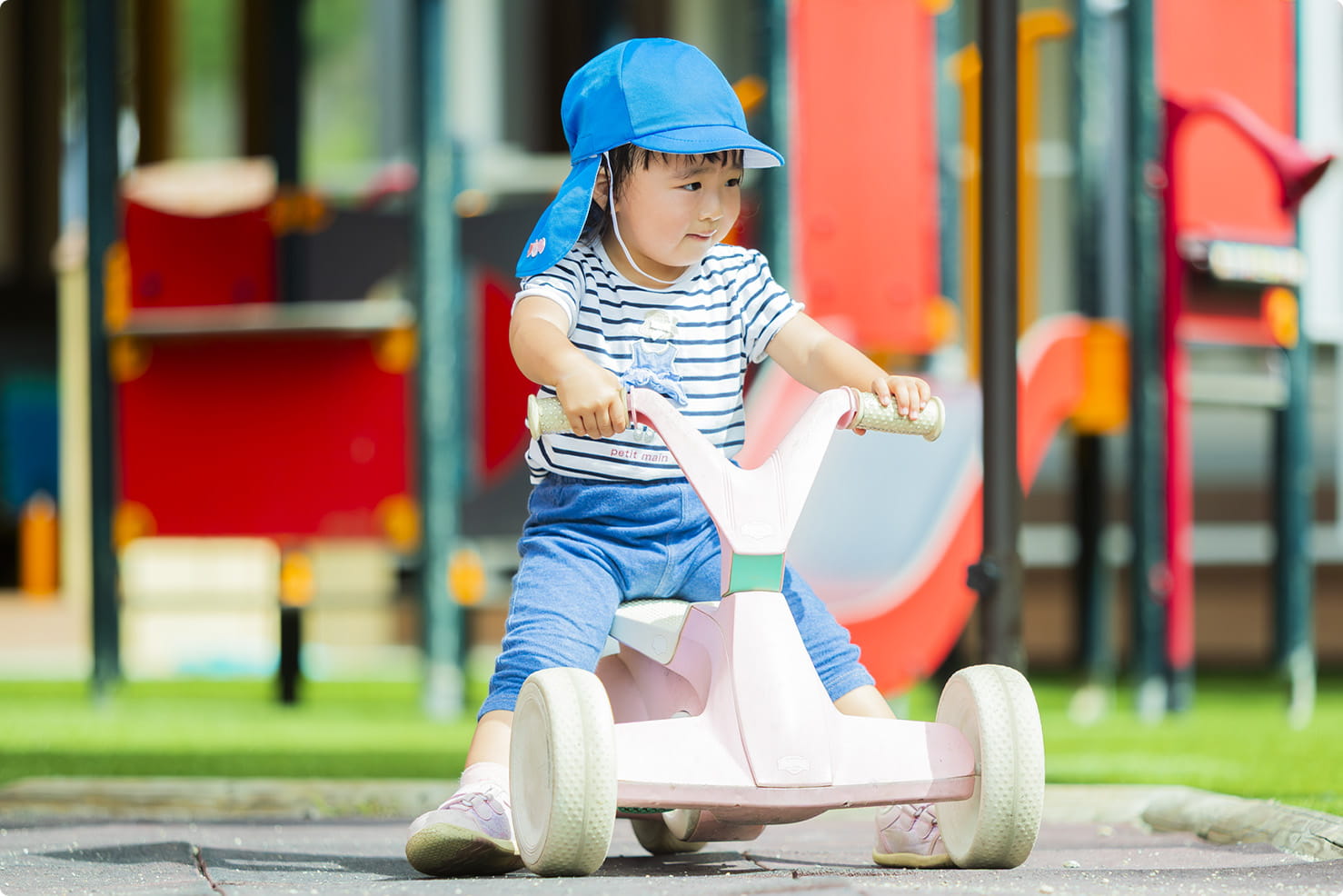 青い帽子をかぶった子どもが三輪車に乗って園庭で遊んでいるシーン