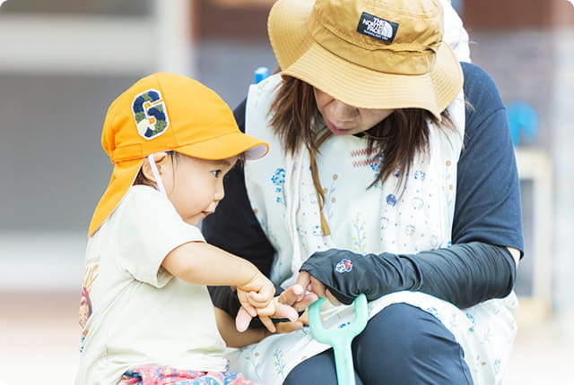 保育士が帽子をかぶった幼児の手を優しく見ている園庭でのやりとり