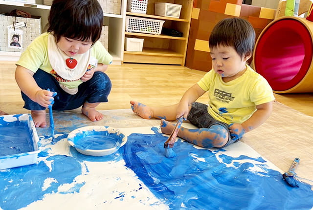 子どもたちが床に広げた紙に青い絵の具で大胆にお絵描きをしている様子