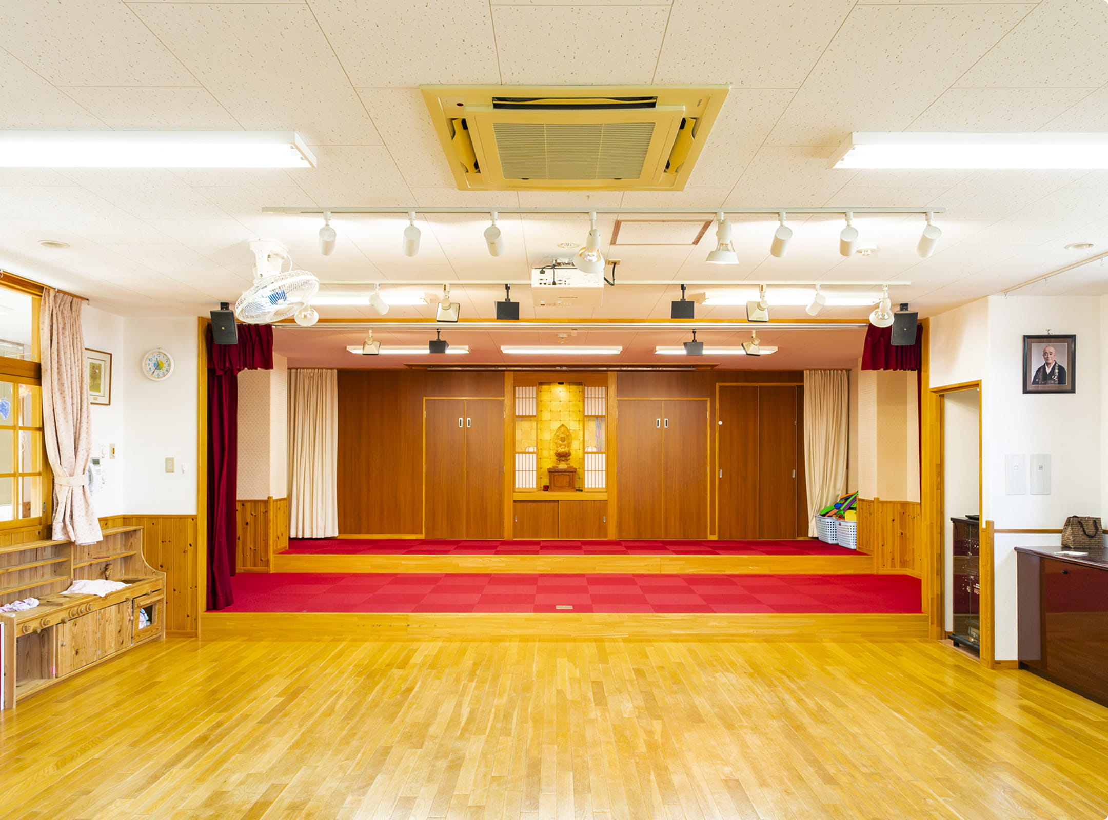 木製のステージと赤いカーペットが特徴的な、保育園内の多目的ホールの風景